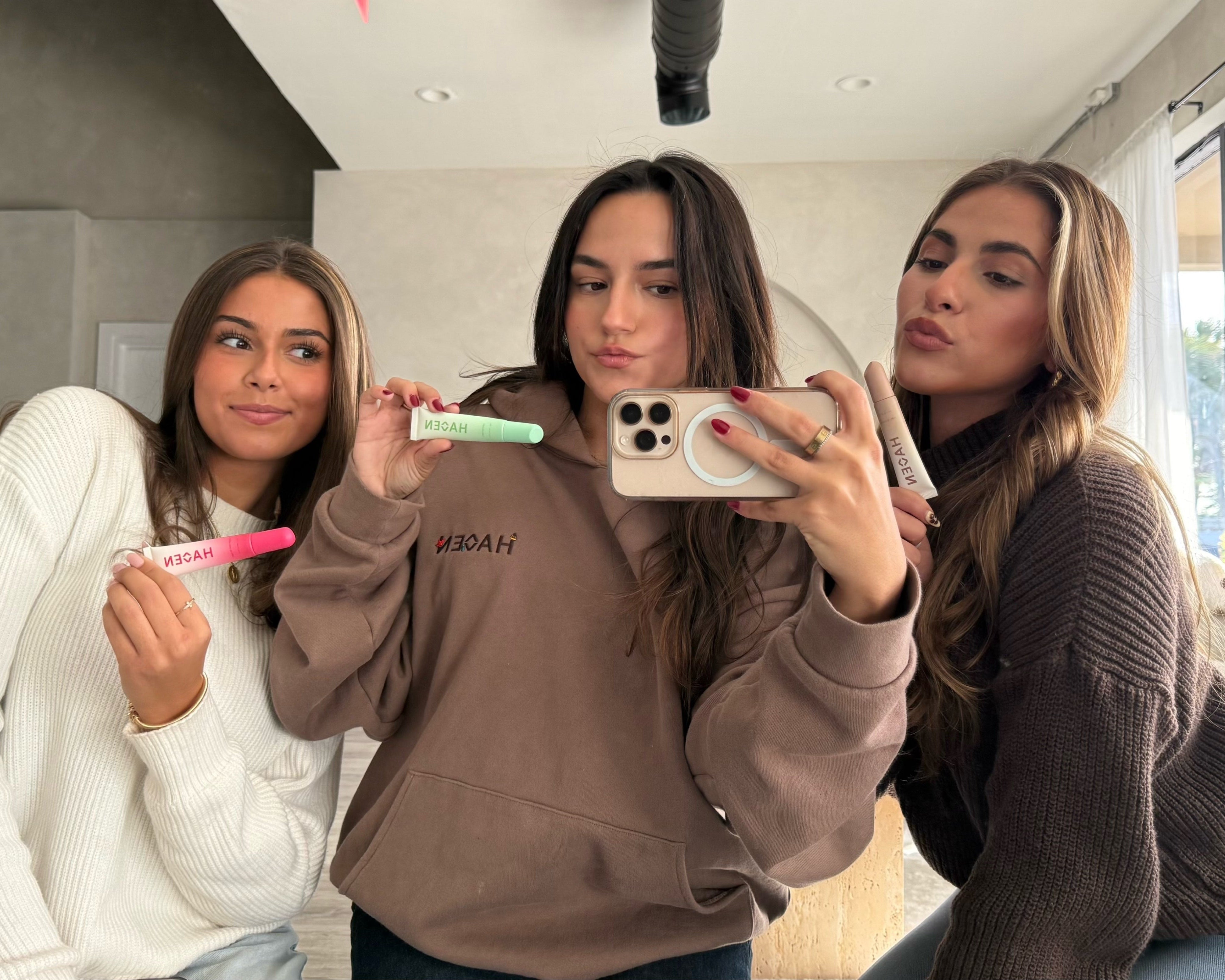 Three women in a modern living room taking a selfie with their lip balms.